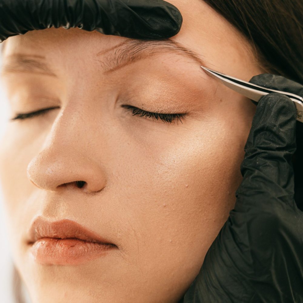 Detailed image of woman getting eyebrows plucked with tweezers by a beautician wearing gloves.
